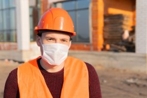 Portrait of male construction worker in medical mask and overalls on background of house under construction.