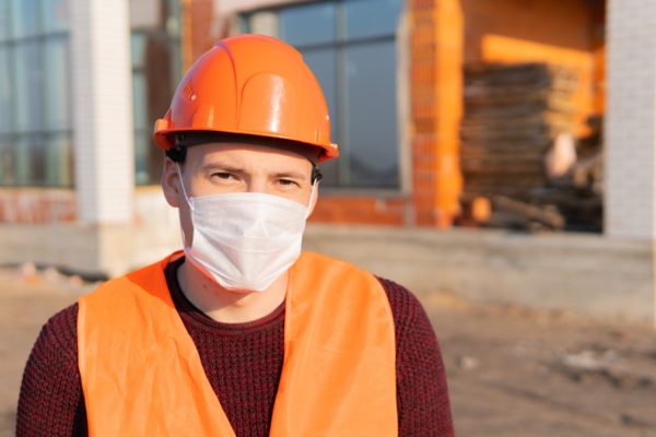 Portrait of male construction worker in medical mask and overalls on background of house under construction.