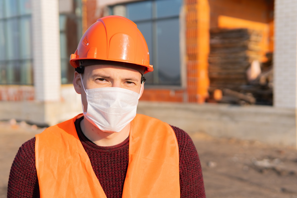 Portrait of male construction worker in medical mask and overalls on background of house under construction.